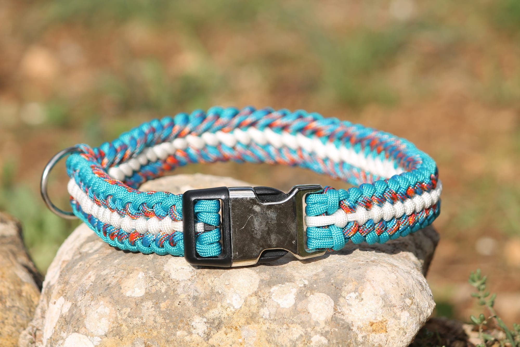 Collier pour chien en paracorde tressée turquoise et blanche mouchetée d'orange, fermoir clip noir et anneau inox, posé sur une pierre.