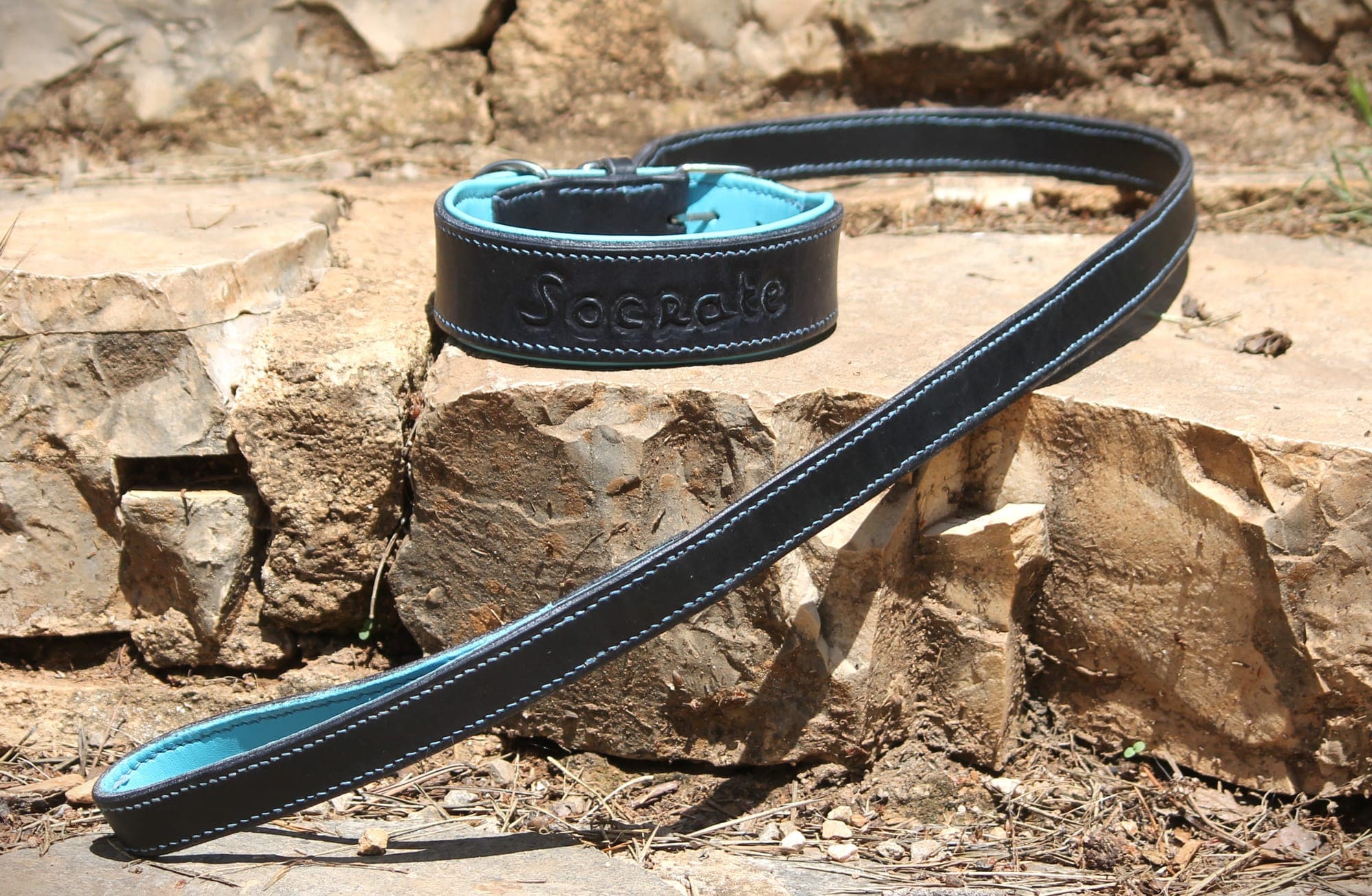 Collier en cuir noir doublé de turquoise, gravé du nom « Socrate », et sa laisse assortie surpiquée de fil bleu, sur un muret de pierre.