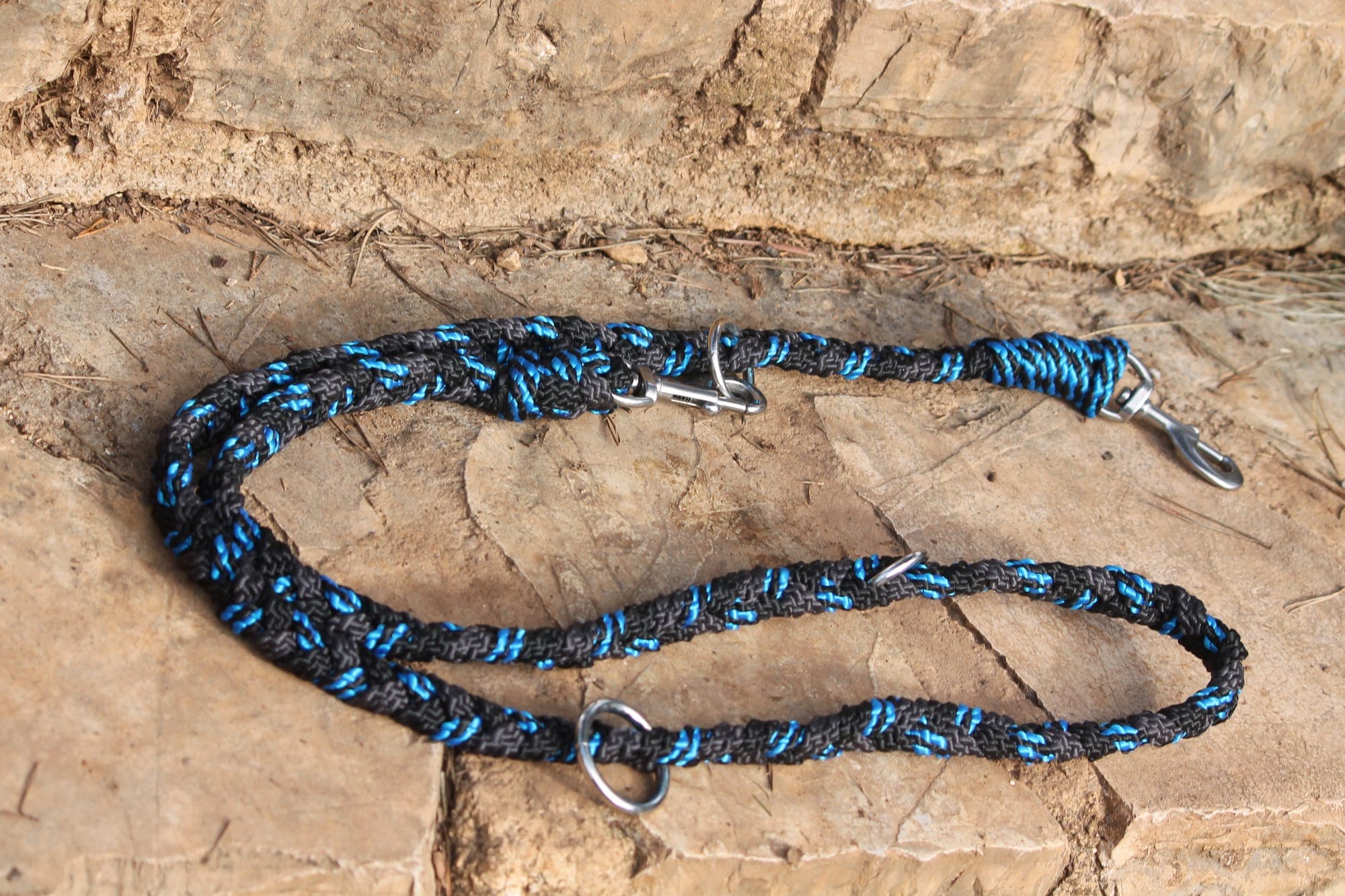 Laisse multipositions en corde tressée noire et bleue, deux mousquetons inox et anneaux de réglage, posée sur une dalle de pierre.